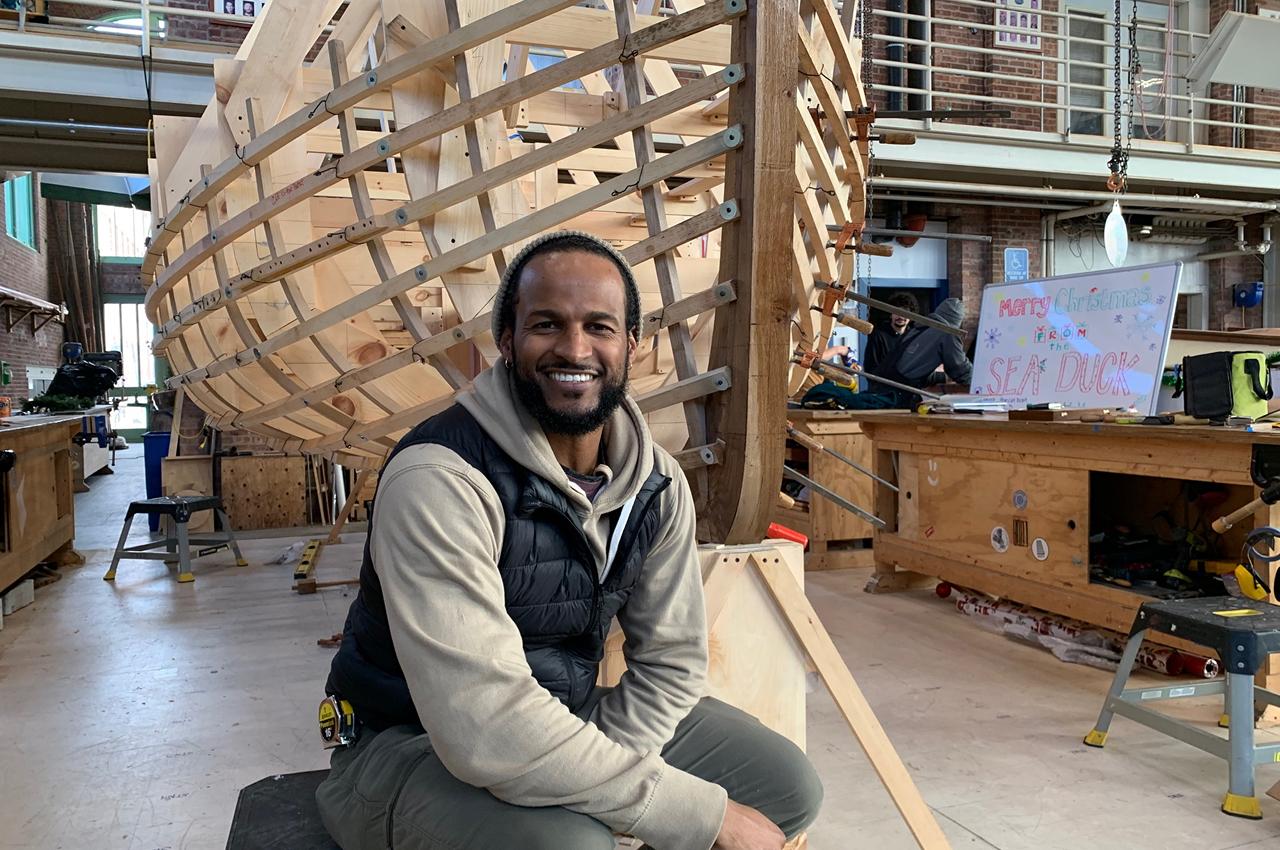 Person sitting in front of a large wooden boat frame under construction.