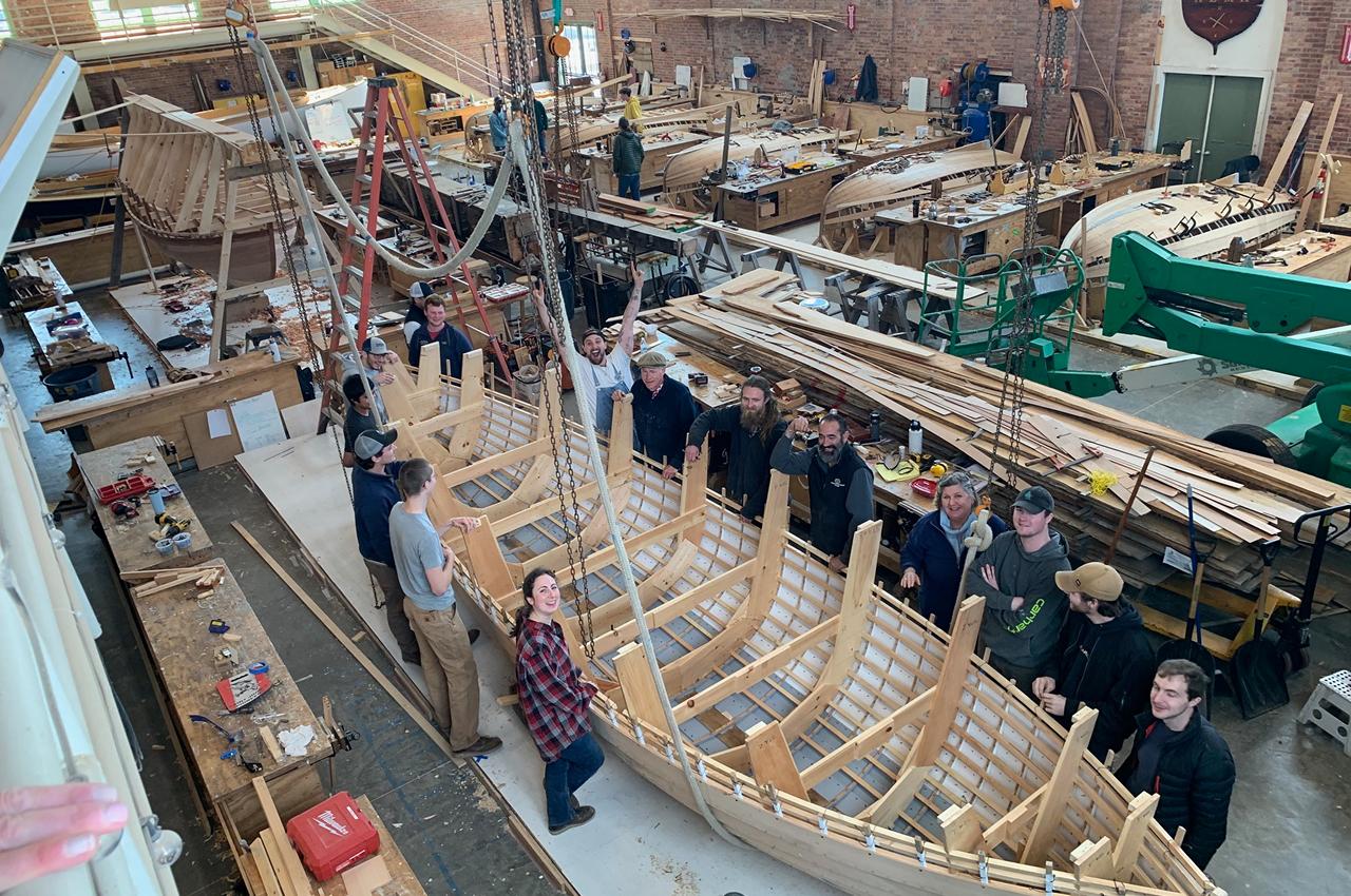 Group of people standing next to a wooden boat under construction, smiling.