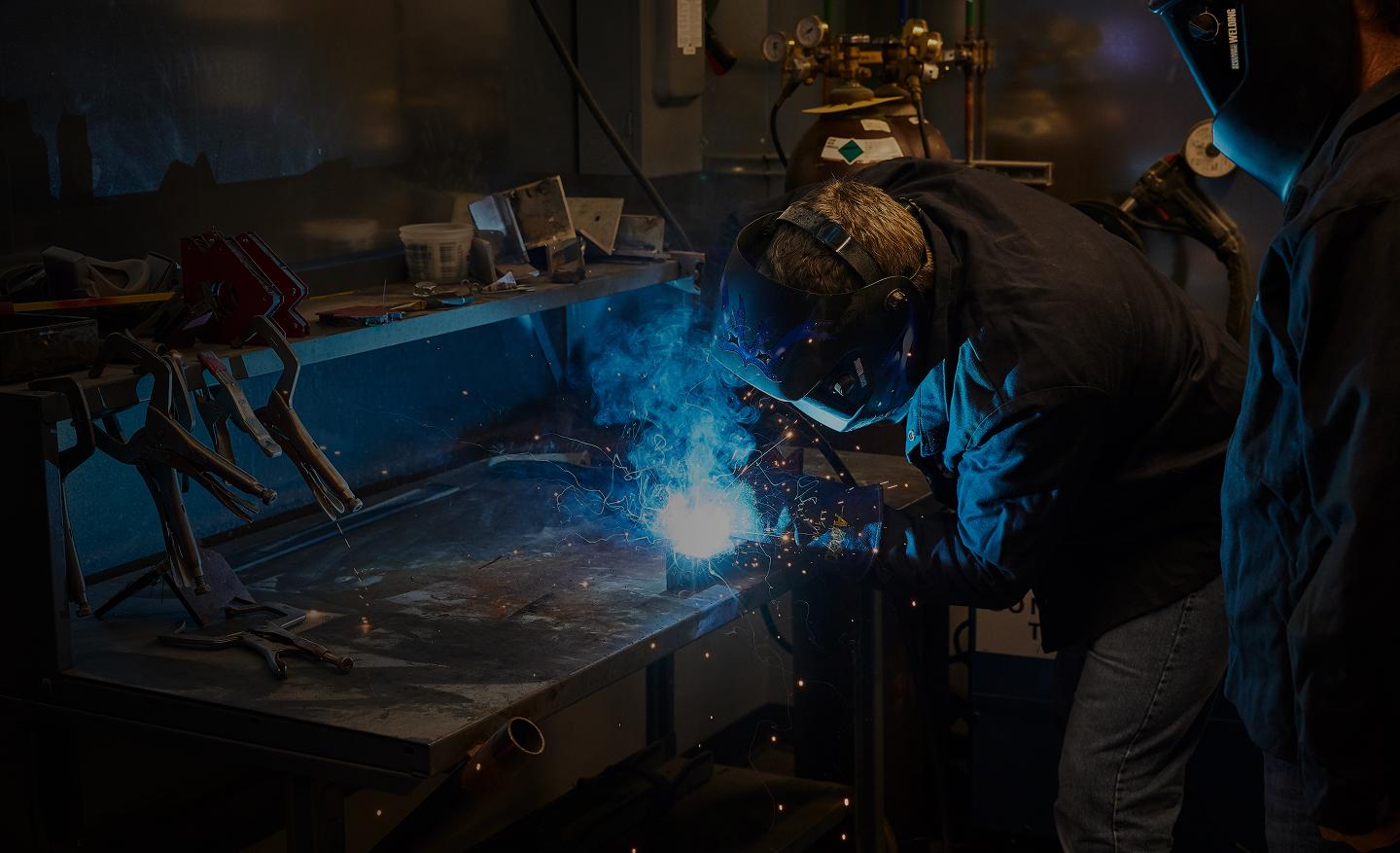 Welder wearing protective gear working on a metal piece with sparks flying.